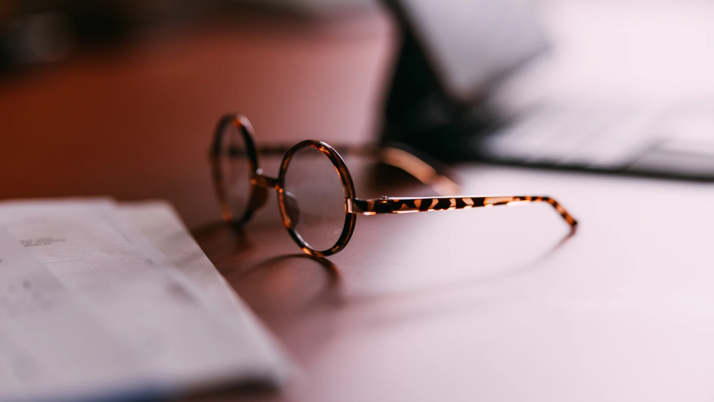 A pair of round, tortoise-shell eyeglasses rests on a desk next to a sheet of paper, with a soft, warm light casting a shallow depth of field.