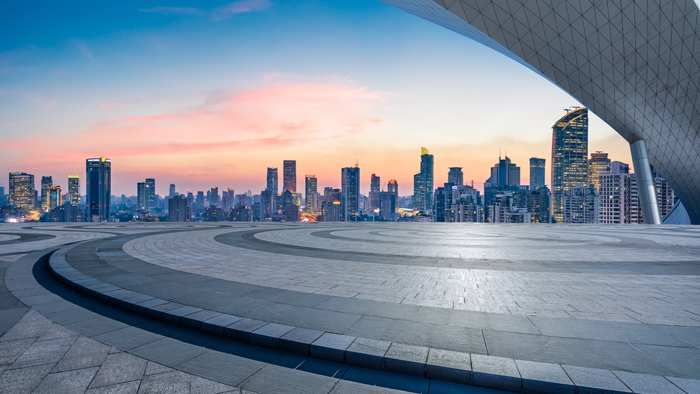 Modern city skyline with high-rises at dusk, viewed from an empty, patterned plaza. A large, curved roof element frames the upper right.